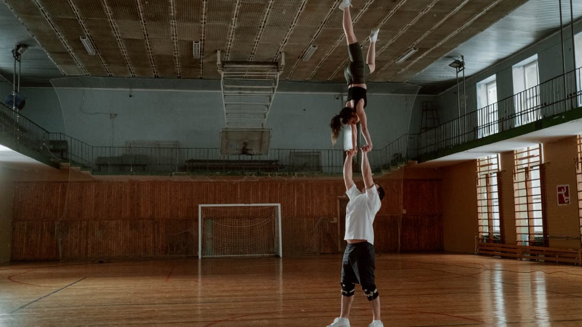 Two people perform an acrobatic balance stunt in a spacious indoor gymnasium.