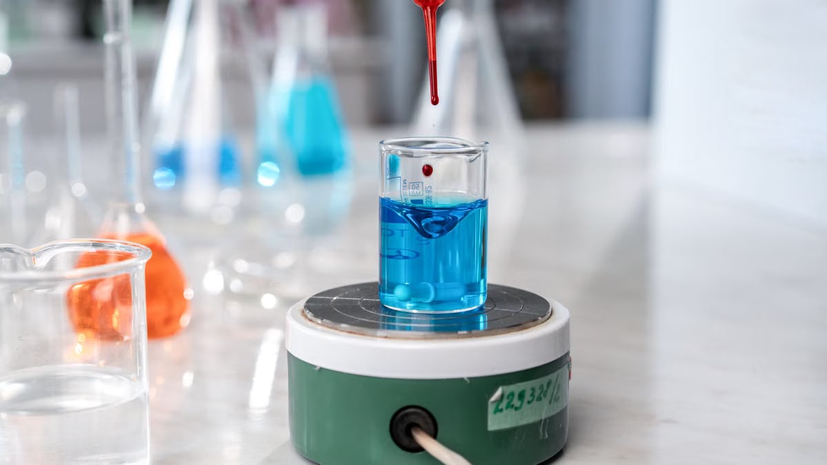 A laboratory experiment with a dropper adding red liquid to blue solution in glassware on a magnetic stirrer.